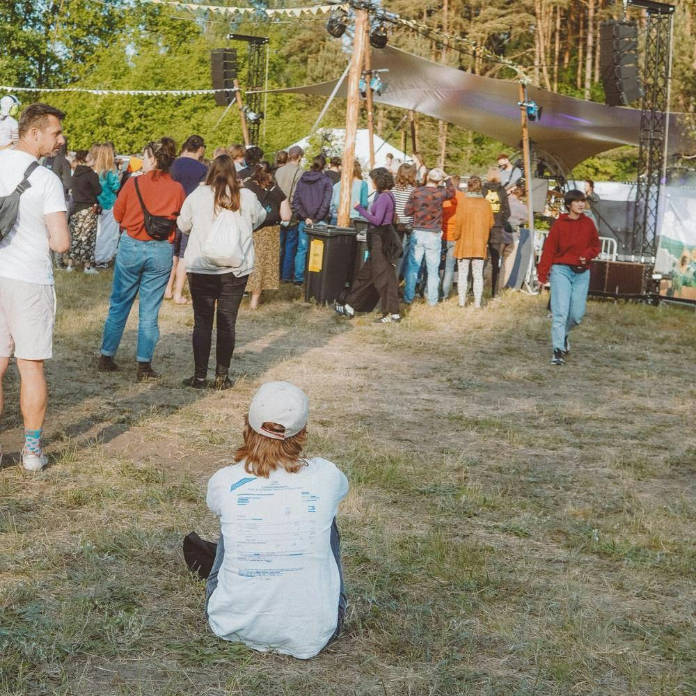 Person auf der Wiese sitzend von hinten mit Blick Richtung Birkenhain-Bühne